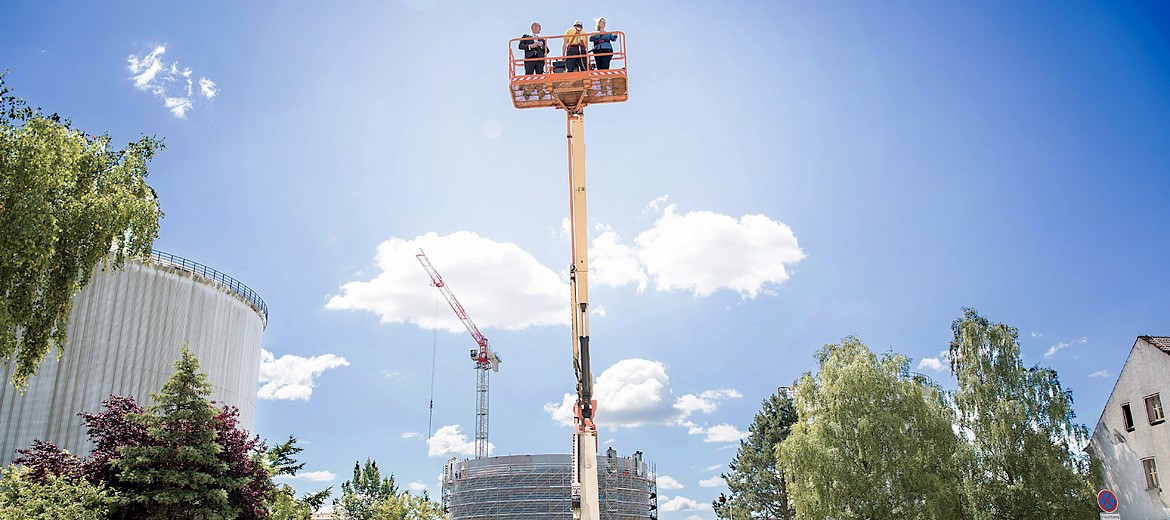 Drei Männer im Steiger vor blauem Himmel, im Hintergrund Gebäude, Bäume und ein Kran auf einer Baustelle
